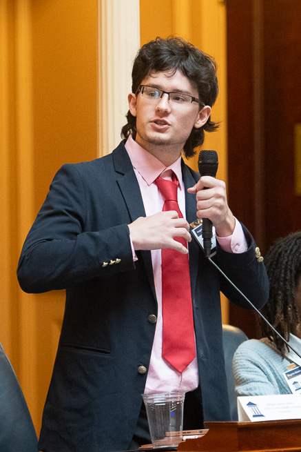 Man wearing a sports jacket and tie speaks into a microphone